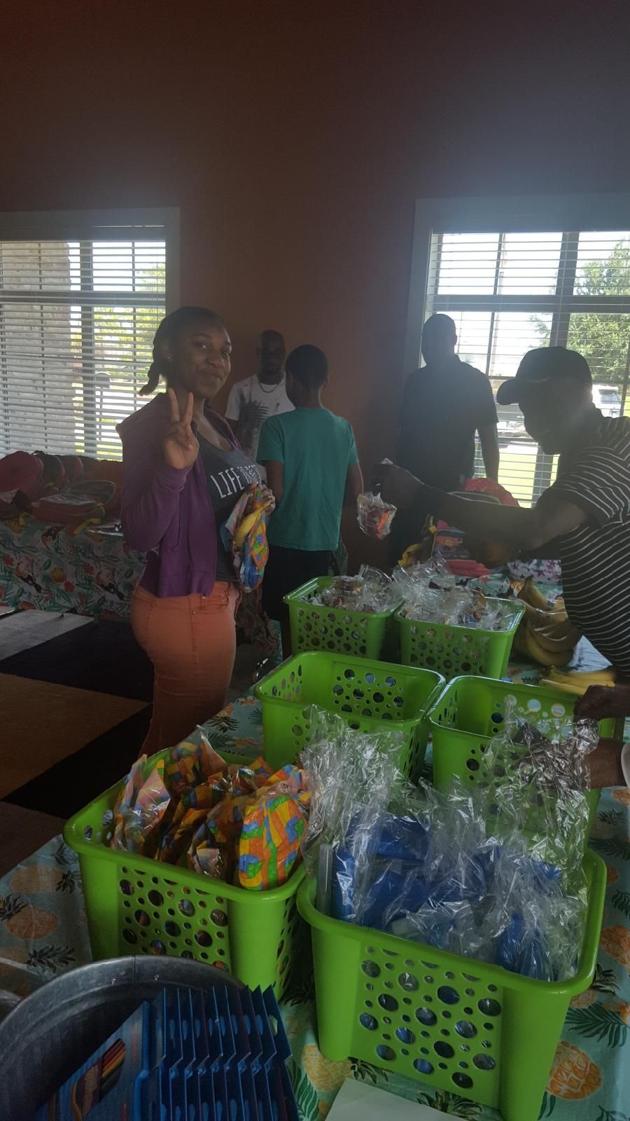People at a table with green baskets of wrapped items. A woman smiles, other people in background. Indoors.