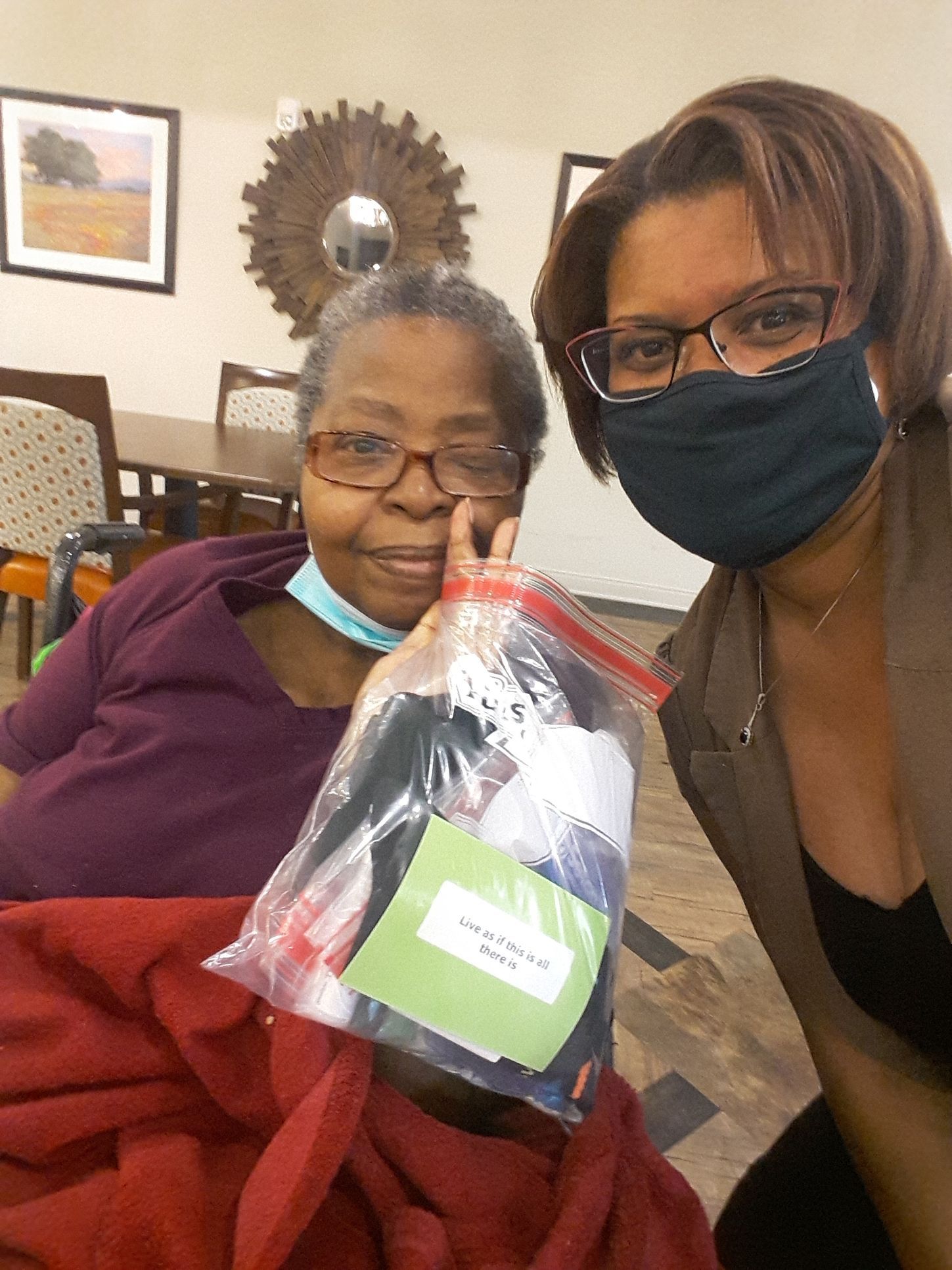 Woman in glasses smiles, holding a bag of supplies next to another woman wearing a mask. Indoor setting.