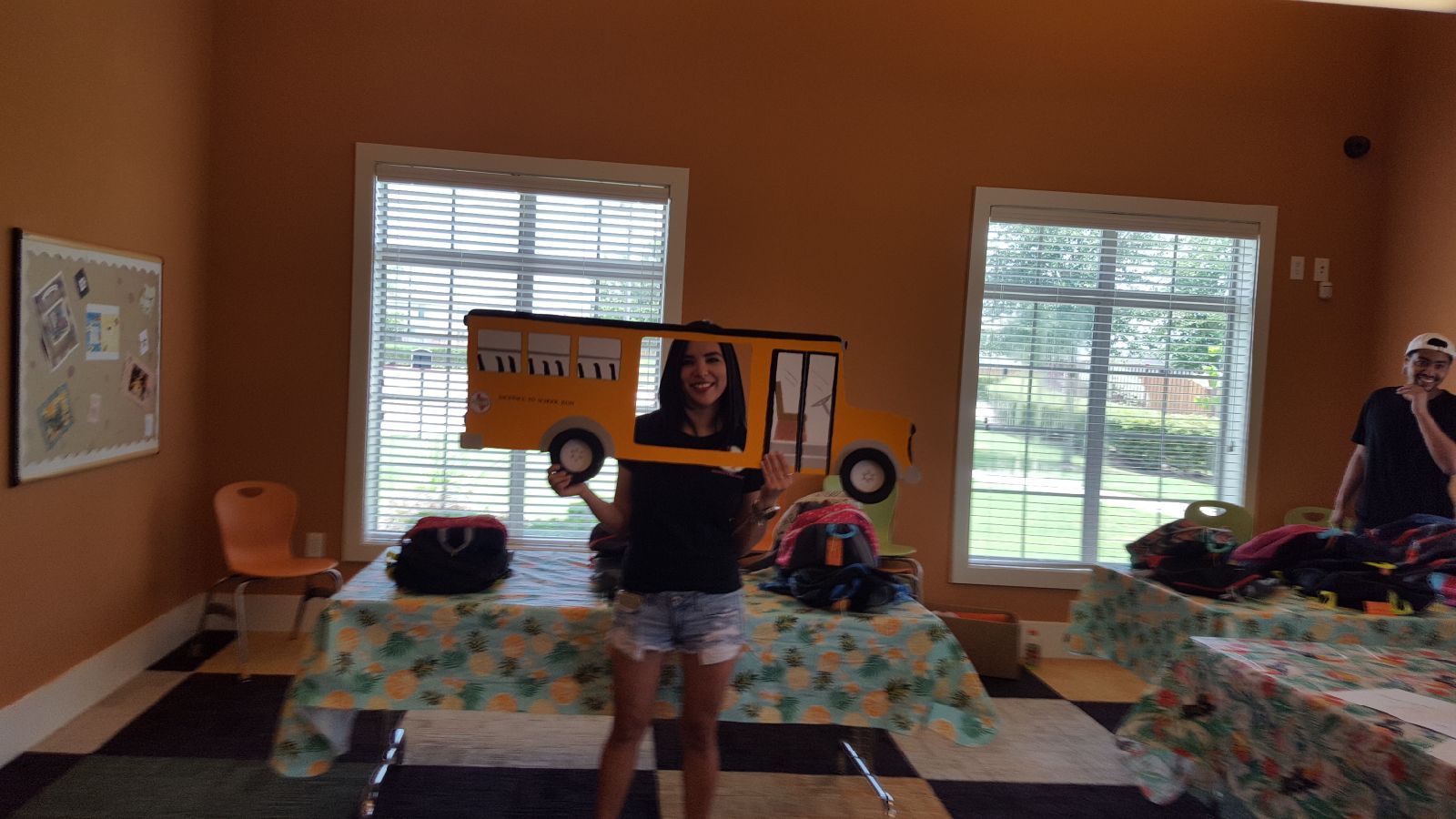 Person holding a yellow school bus cutout, standing in a room with windows and tables.