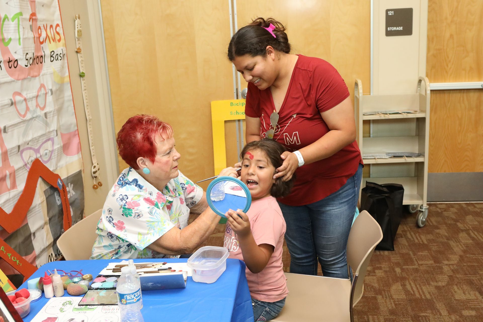 Woman paints child's face at a table, another woman watches. Event in a room with a banner.