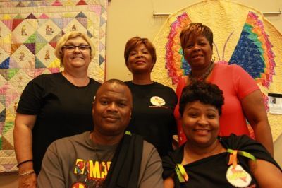 Group of people posing in front of colorful quilts, smiling. Some wear orange ribbons/pins.