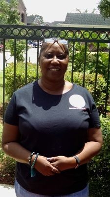 Woman in black shirt, smiling, standing in front of a decorative fence and greenery on a sunny day.