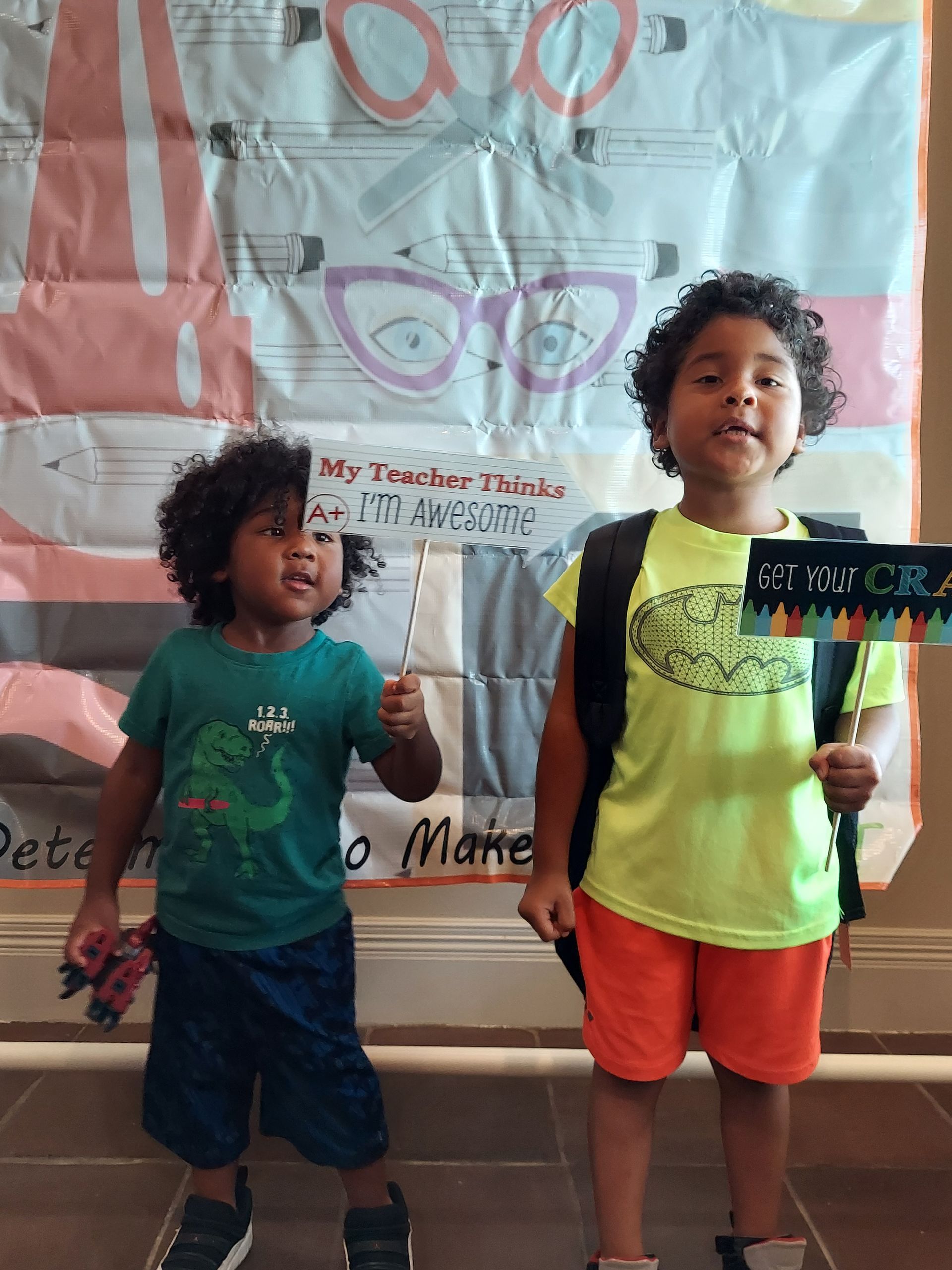 Two children stand in front of a colorful backdrop. One holds a sign saying 