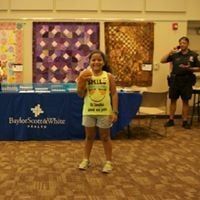 Girl holding food smiles at camera; Baylor Scott & White Health banner, quilts in background.