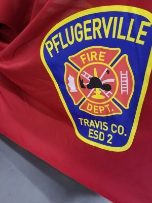 Pflugerville Fire Dept. logo on red fabric. Blue and yellow badge with fire tools and Travis County info.