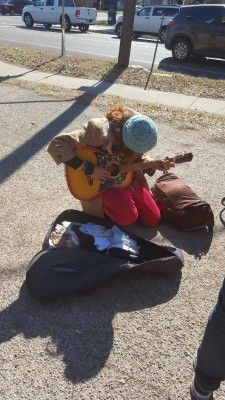 Person playing guitar outdoors, seated on pavement next to an open case, wearing hat and coat.