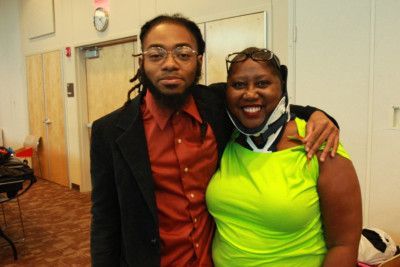 A person with dreadlocks and a person in a green top smile, arms around each other, near a door.
