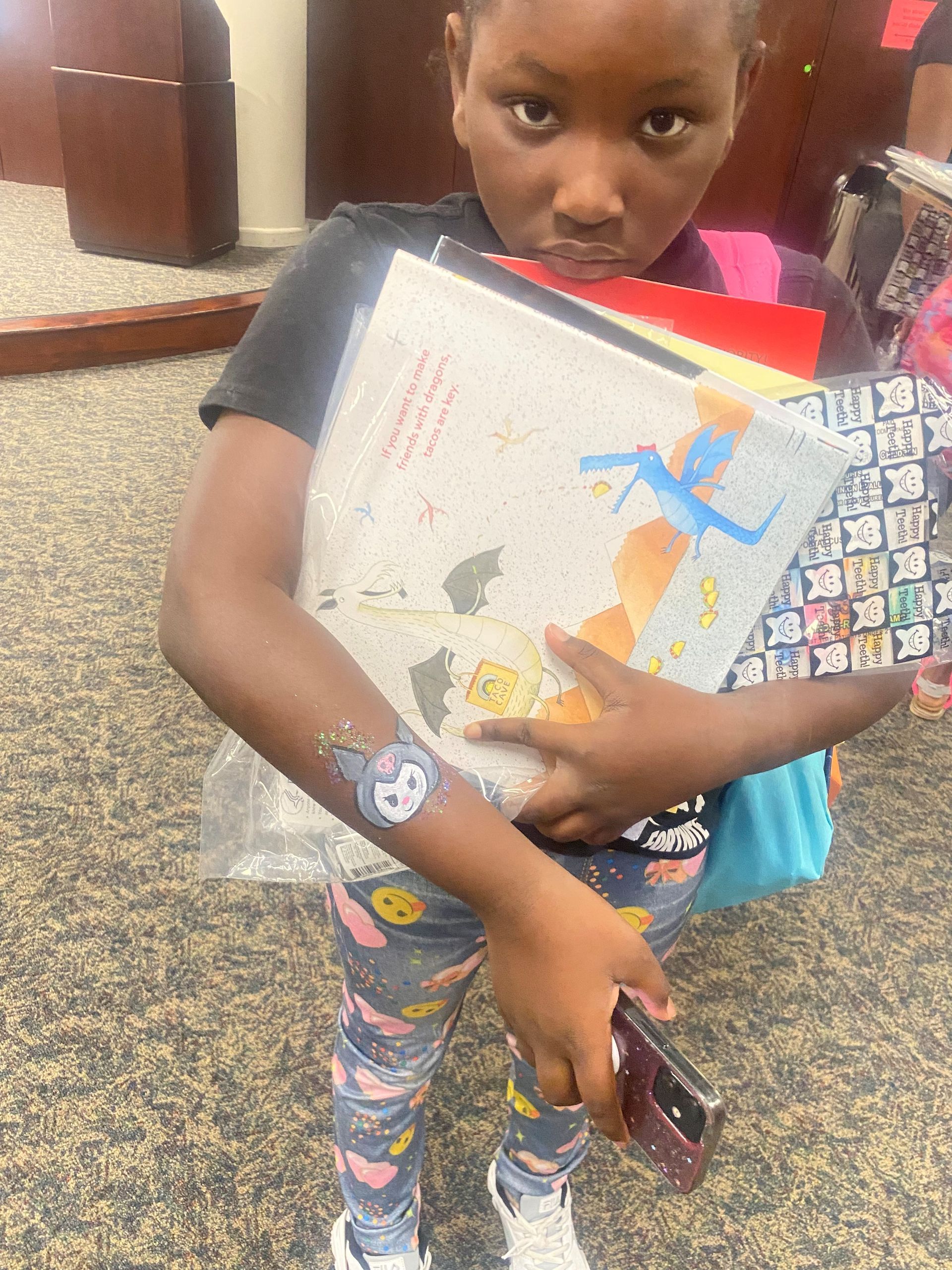 Child holding books, looking at the camera. They wear patterned pants, have a temporary tattoo, and a phone. Indoors.