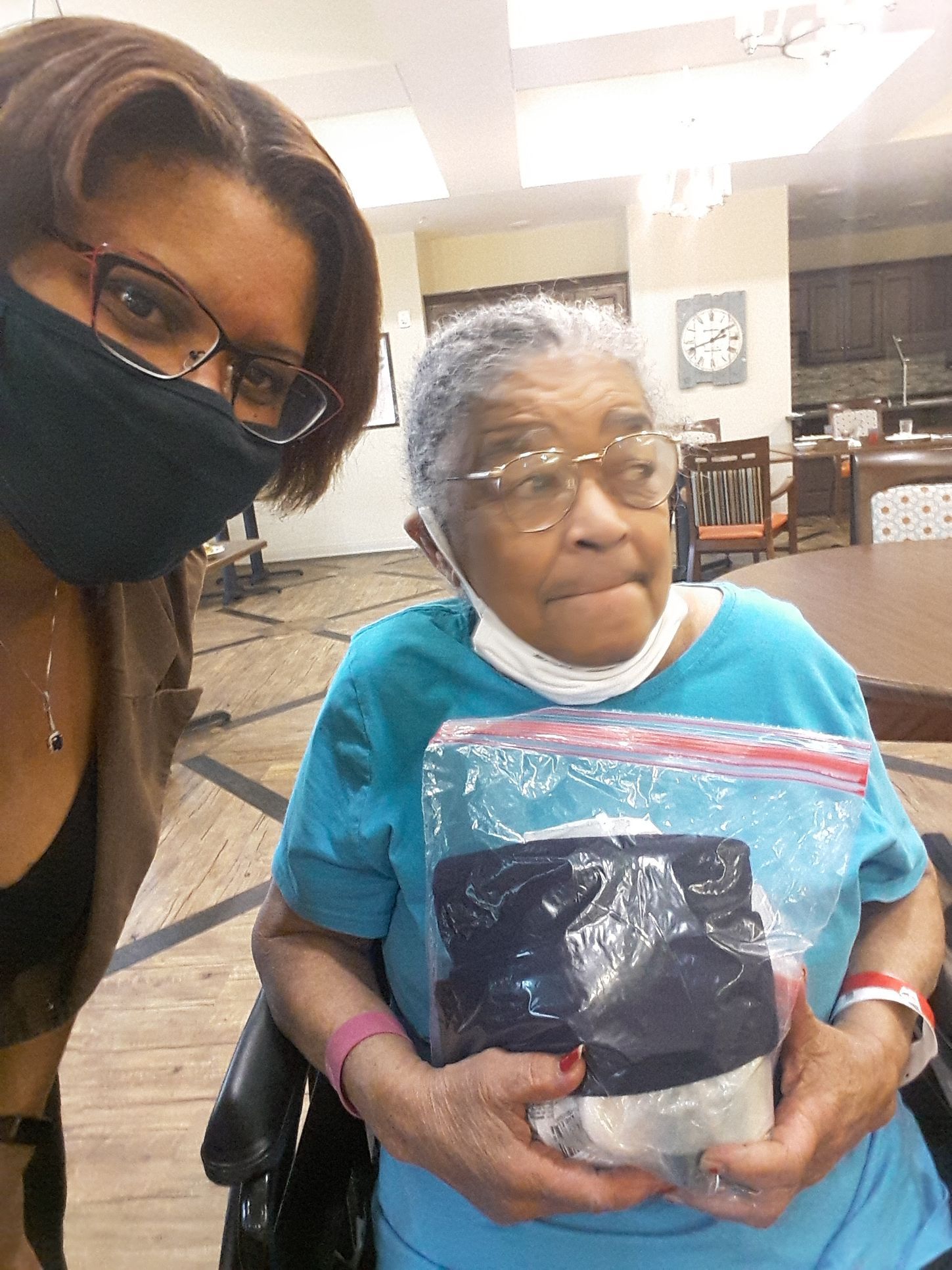 Woman in mask with older woman in wheelchair, holding package. Interior, wooden table.