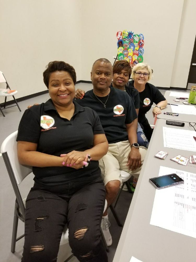 Four people sitting at a table. They all wear black shirts with logos, smiling. Indoors, bright lighting.