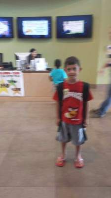 Boy in Angry Birds shirt stands indoors, smiling, near a counter with screens above.