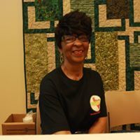 Woman with glasses and short hair smiles, wearing a black shirt, seated in front of a patterned wall hanging.