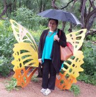 Woman holding umbrella stands near a large butterfly sculpture outdoors; green trees in the background.