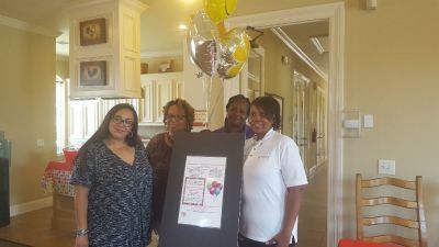 Four people stand indoors, holding a framed poster, with balloons.