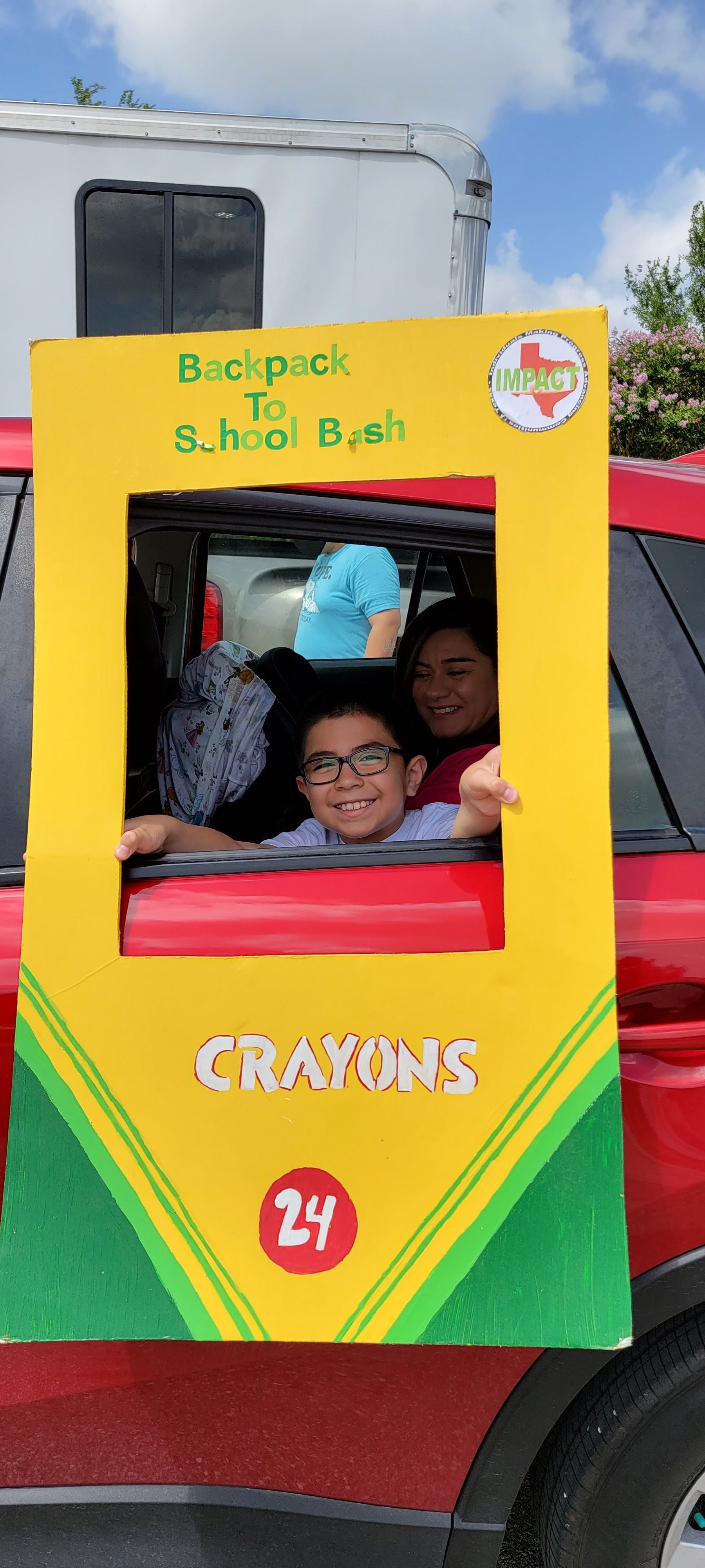 A child and adult pose in a frame shaped like a crayon box. The frame is attached to a red car during a 