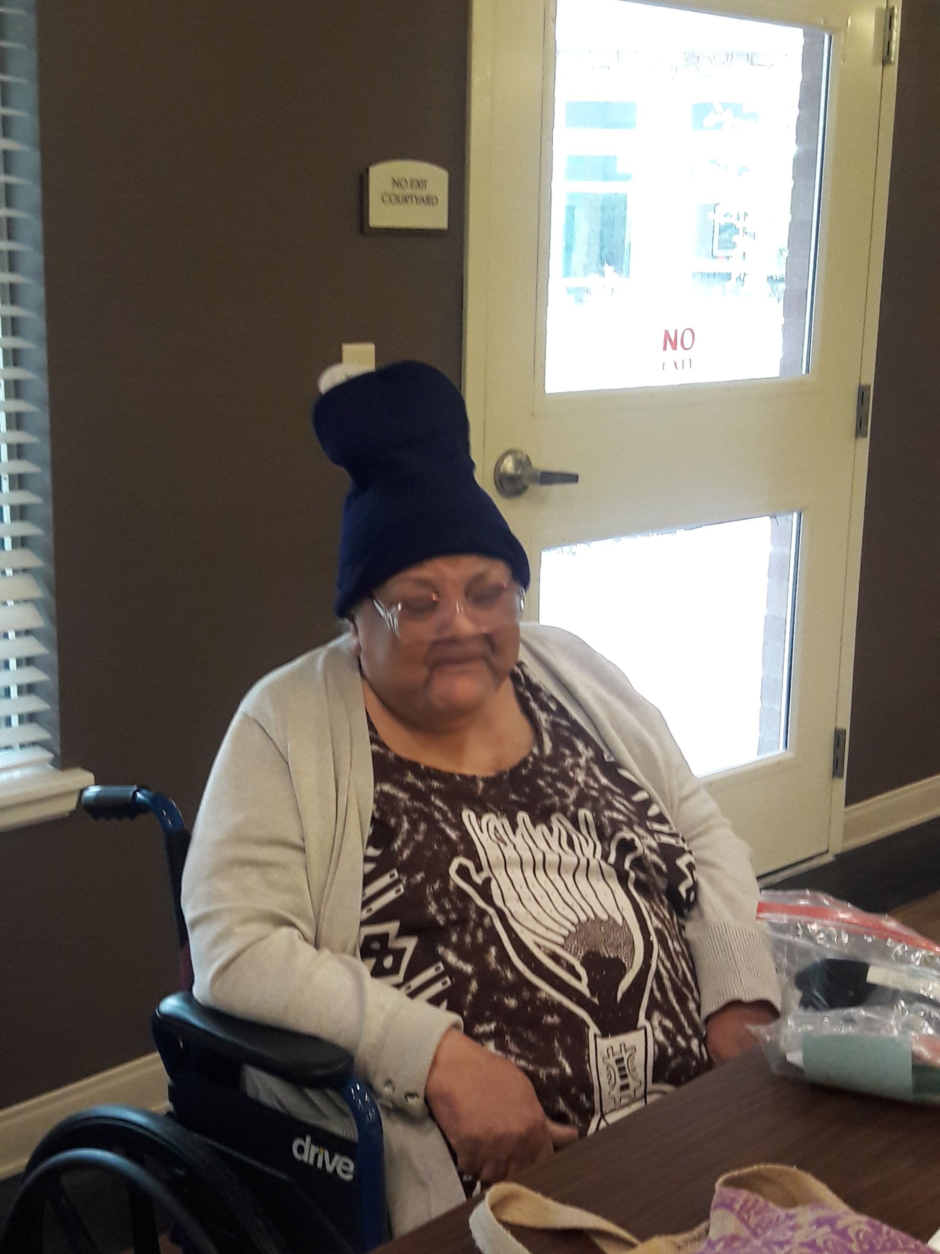 Woman in wheelchair wearing a dark blue beanie and glasses, seated indoors, near a door.