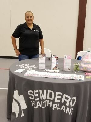 Woman at Sendero Health Plans table, smiling. Gray tablecloth with logo, promotional materials, indoor setting.