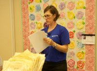 Woman in blue shirt holding papers, standing in front of a colorful quilt.