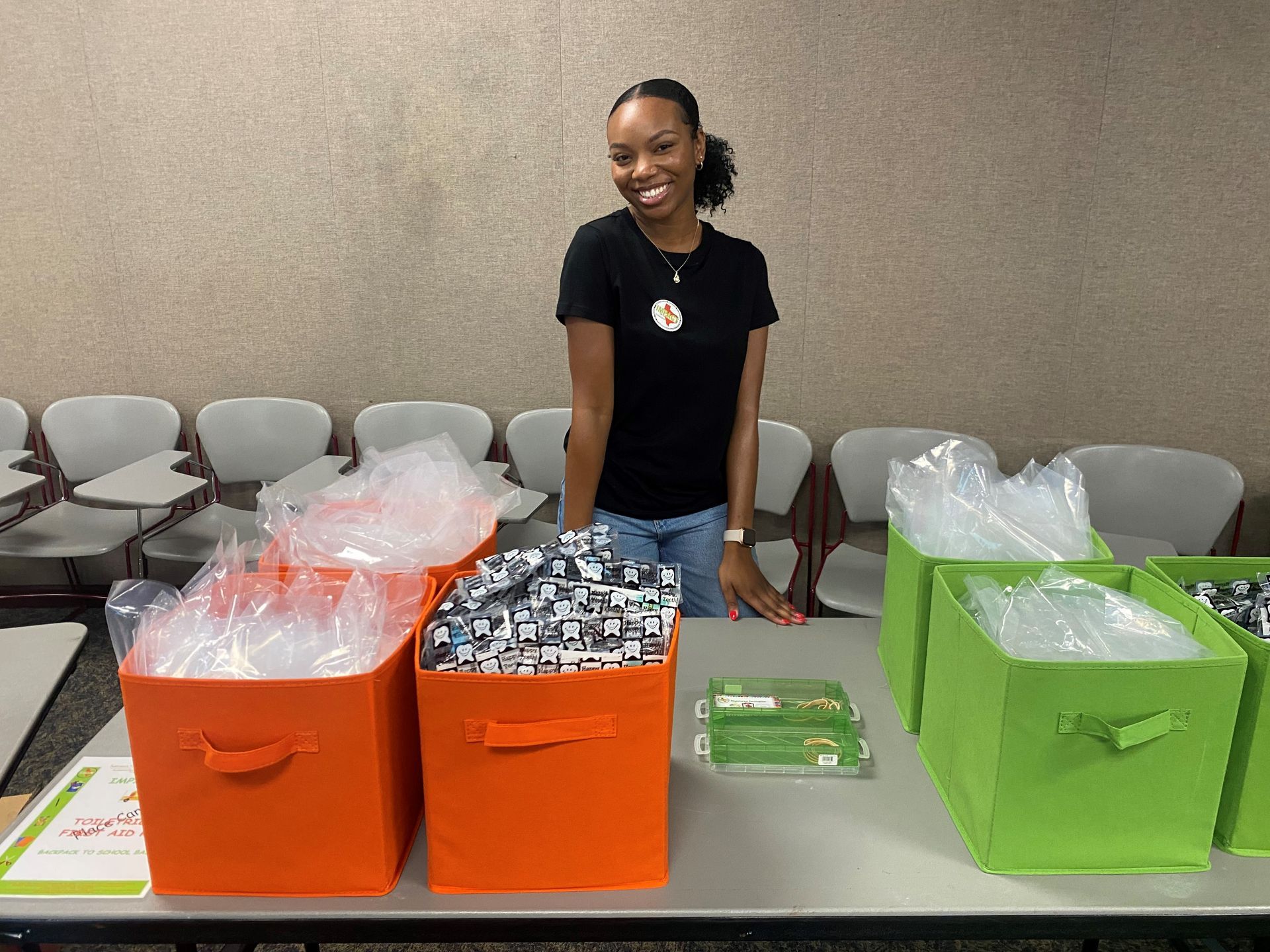 Woman smiles behind a table with orange and green bins filled with wrapped items.