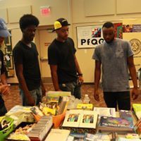 Three people looking at items on a table. The table has various objects, some in an orange container.