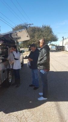 Three people standing near a food truck. One person is handling food, while the others observe. Blue sky in the background.