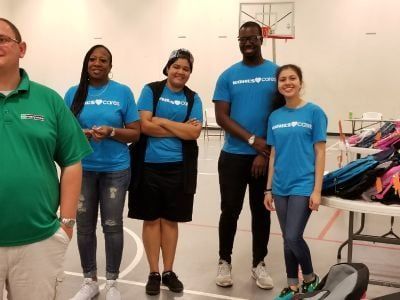 Group of people in blue shirts standing in a gym, posing. Backpacks on table in the background.