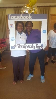 Two people pose behind a Facebook photo frame at Peninsula Park. One wears a white shirt, the other purple. Yellow balloons are above.