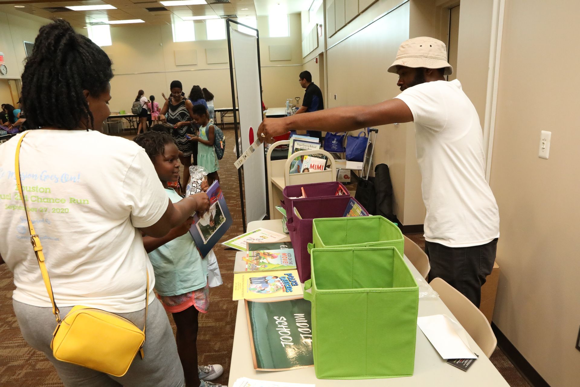 People at a table with books and bins; a man assists a woman and child selecting books.