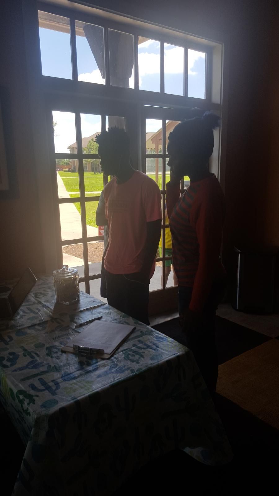 Two people looking out a window. One in a pink shirt, the other in red. Sunny outdoor view.