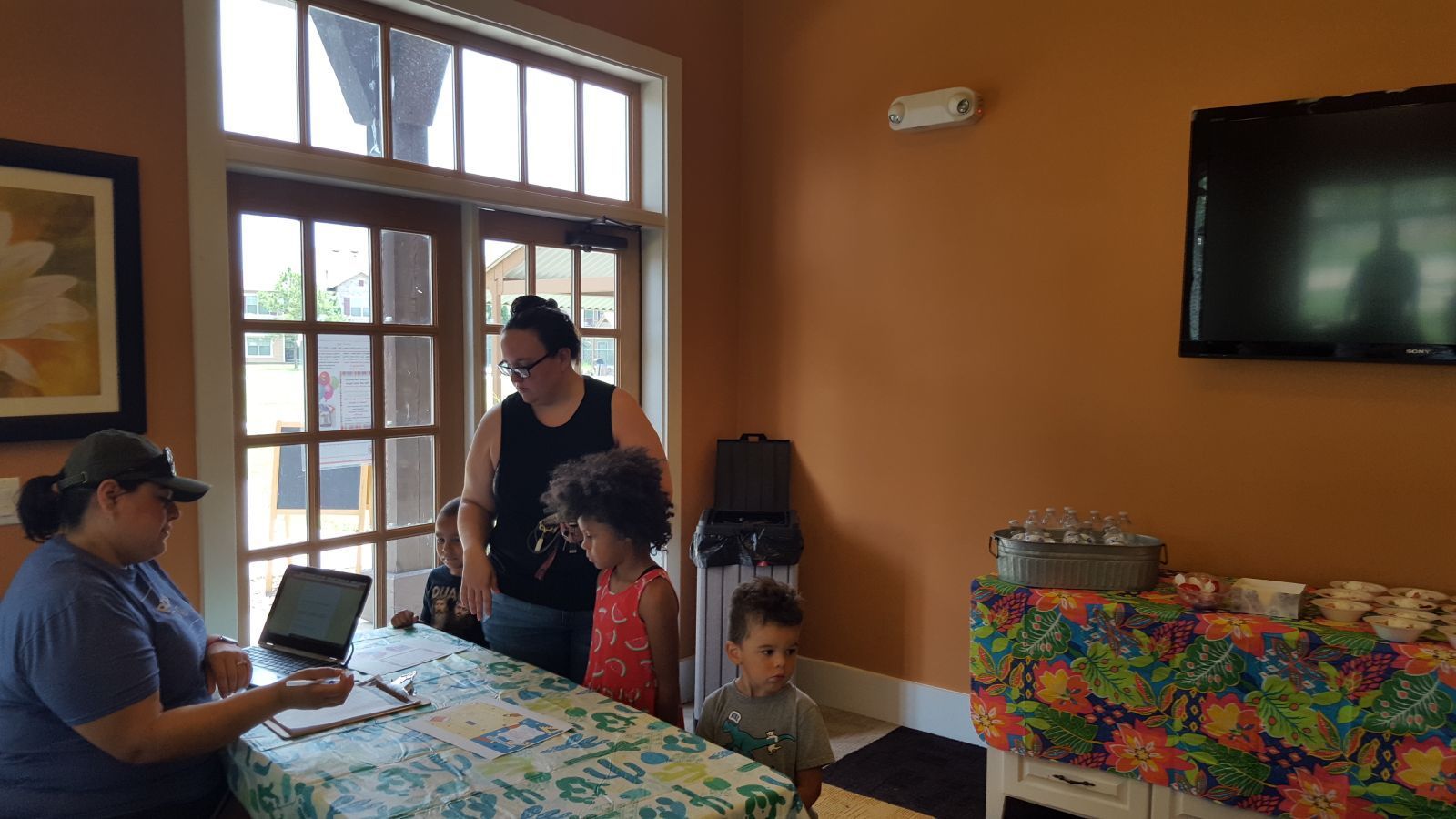 People at a table inside. Woman at left writes on a clipboard. Two children stand near a woman by a door. A TV is on the wall.