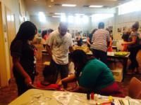 People crafting at a table with supplies. Others observe in a brightly lit room.