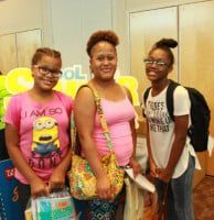 Three people smiling, holding bags and supplies, in front of a school banner.