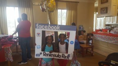 Two children pose inside a Facebook-themed photo frame at a party, other people in the background.