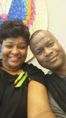 Smiling couple in front of a colorful, abstract art piece. Woman in black top with green accent, man in grey.