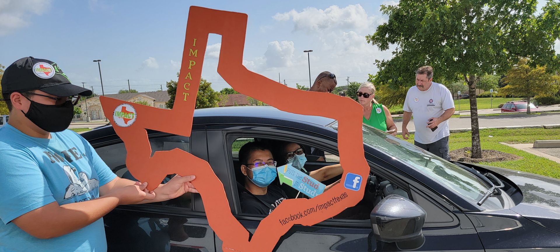 People holding a Texas-shaped cutout greet people in a car. All are wearing face masks. The setting appears to be outdoors.