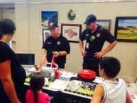 Firefighters demonstrating safety at a table to children and adults indoors with art on the wall.