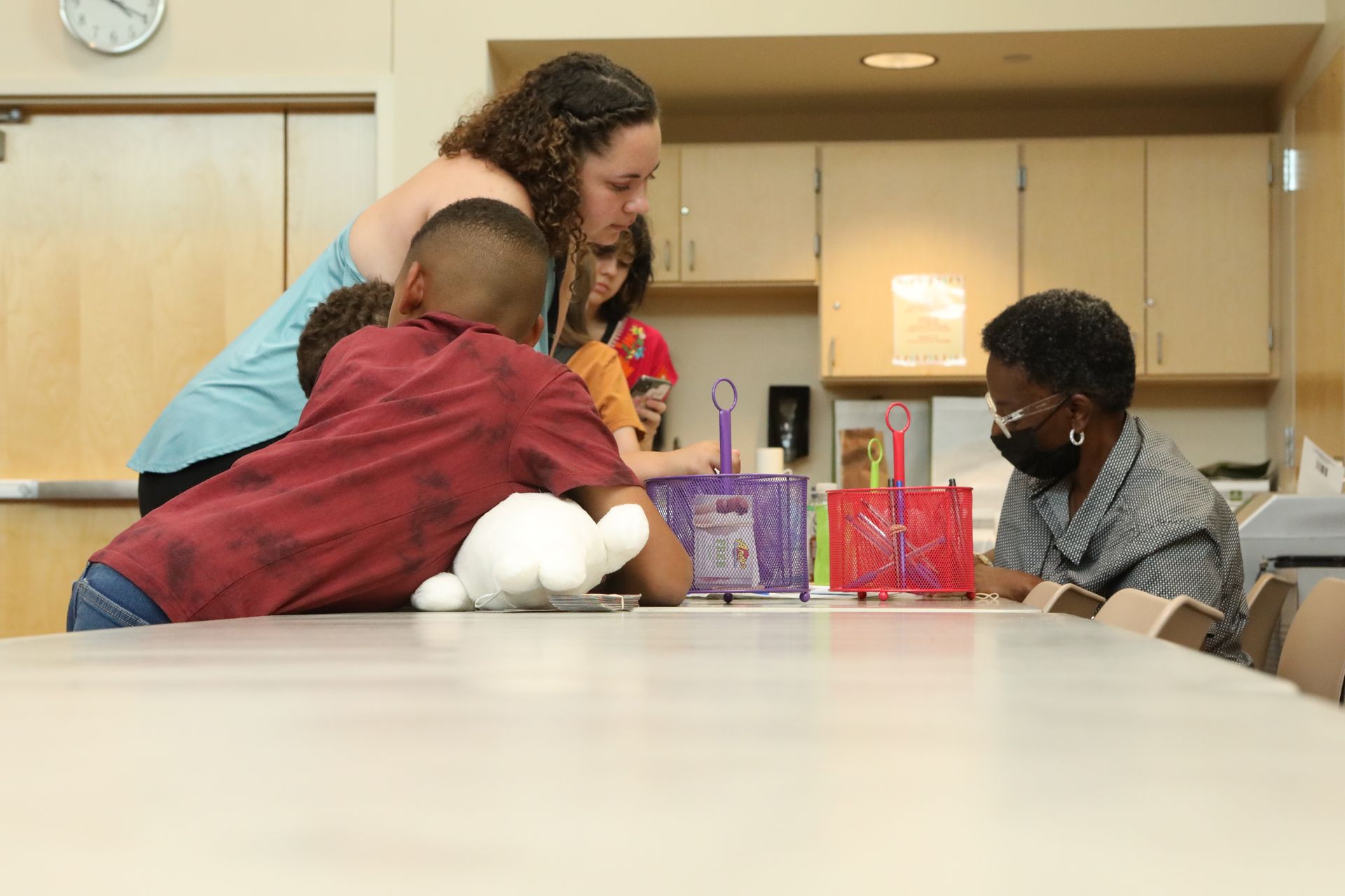People gather around a table with craft bags. An adult assists children with a project in a brightly lit room.