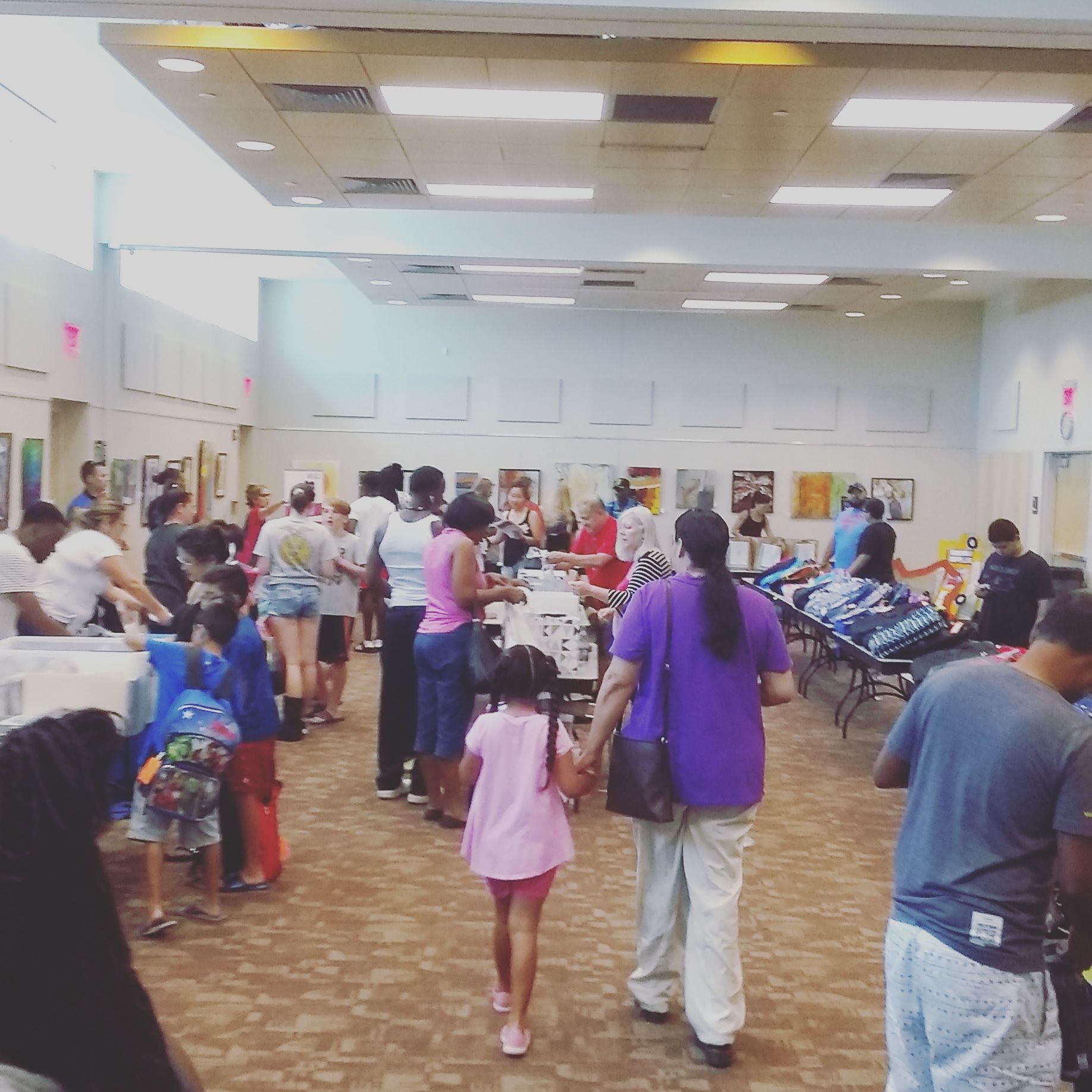 People at an event in a large room, looking at tables and paintings on the walls.