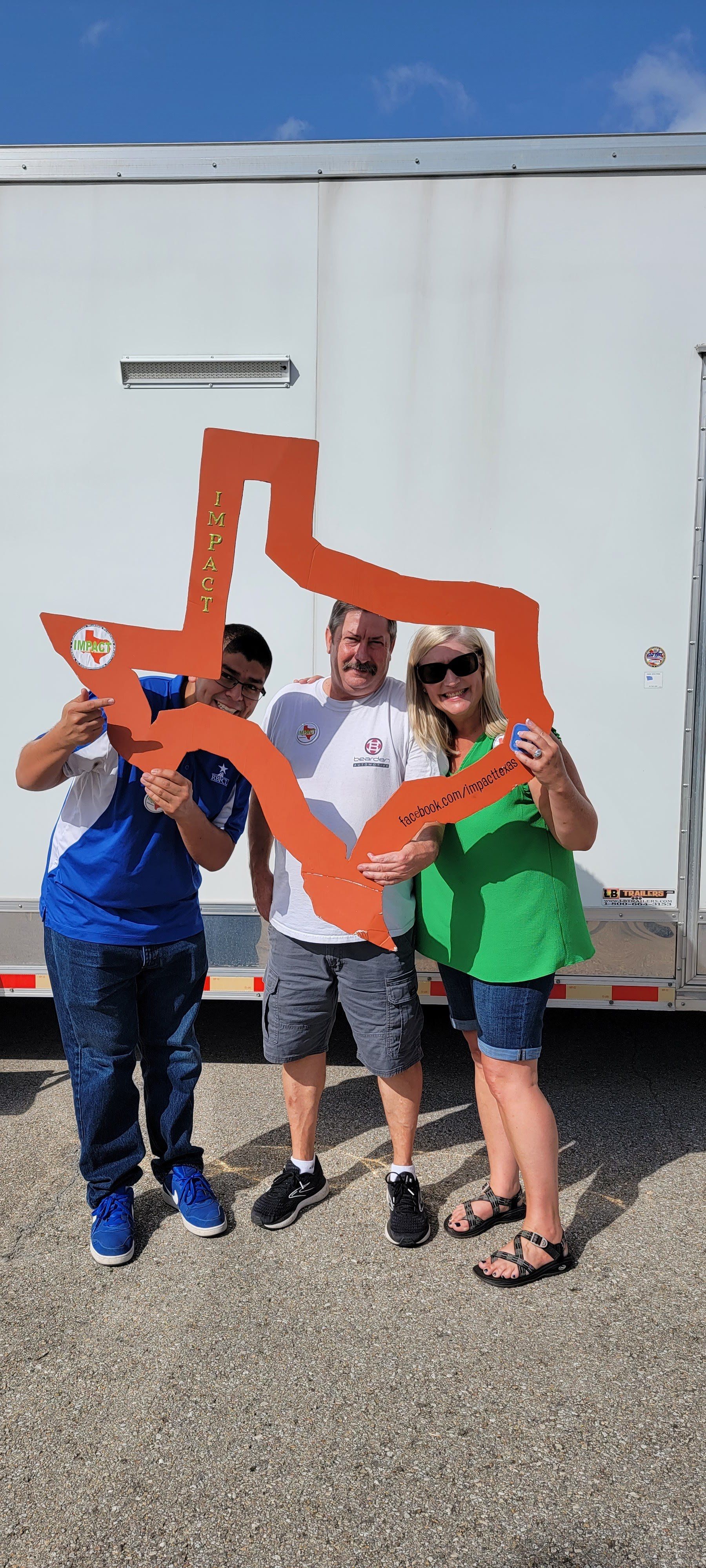 Three people pose with a cutout of the Texas shape. They stand outside near a white trailer.