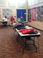 People at tables, backpacks on a table, quilts on wall.