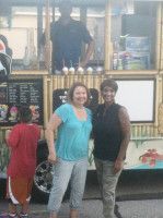 Two women pose in front of a food truck with a person standing inside.