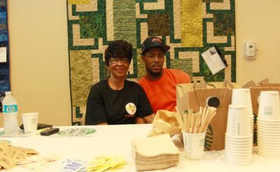 Two people behind a table, smiling. A colorful quilt is on the wall behind them. There are supplies on the table.