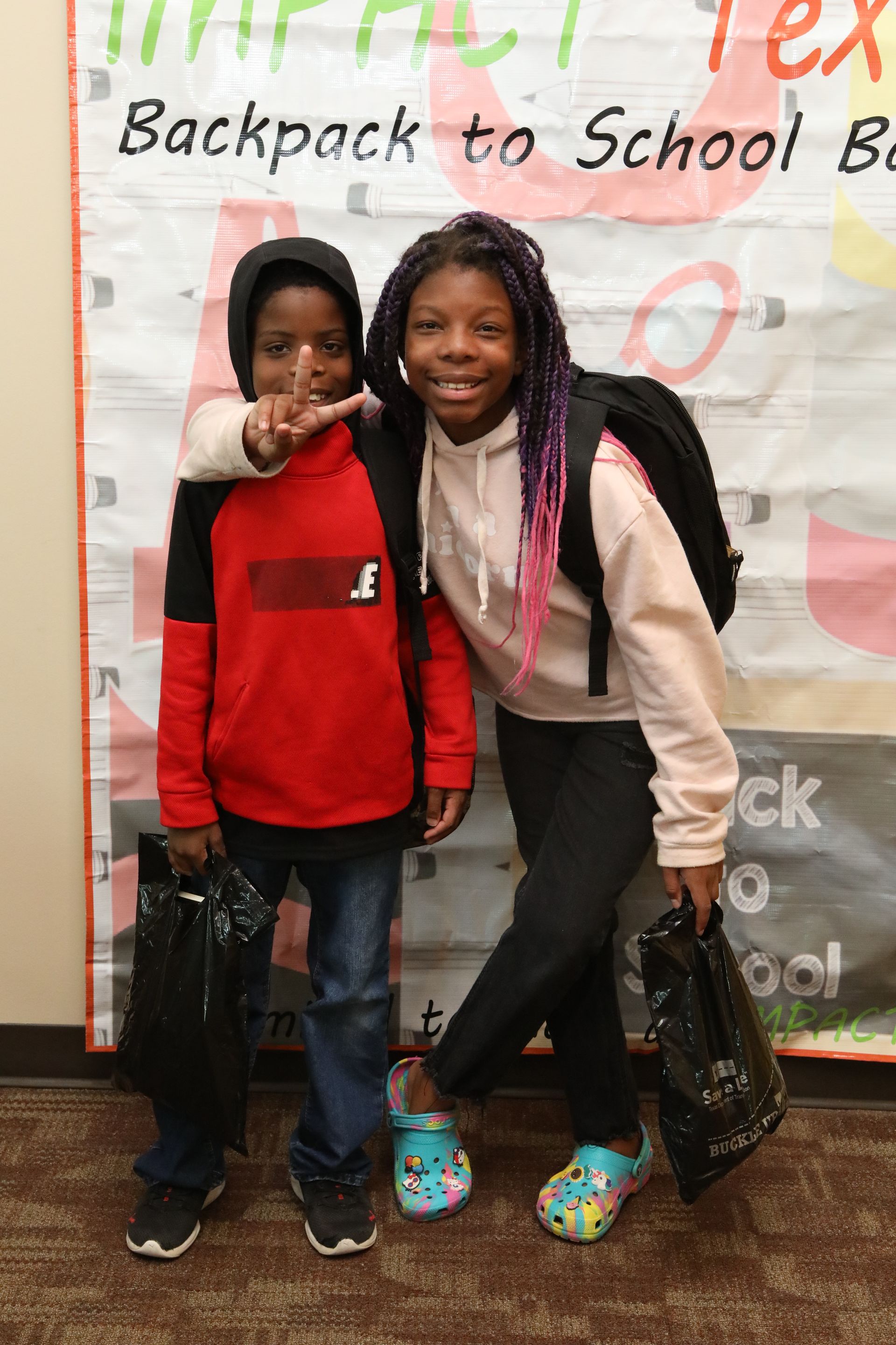 Two children pose with backpacks and bags for a back-to-school event; one child gives a peace sign.