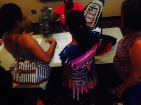 People at a table looking at backpacks. One person in pink shirt hands someone a bag.