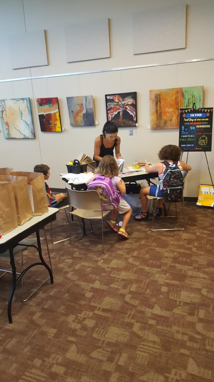 Kids at a table with an adult. Brown carpet. Artwork on wall.