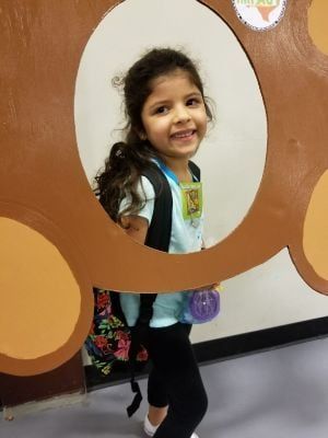 Young girl smiles, framed by a cutout, wearing backpack and carrying a purple item.