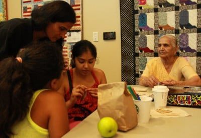 People seated around a table. A woman and two young girls work with paper. An older woman watches them. A paper bag and a cup are on the table.