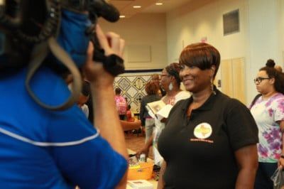 Woman in black polo shirt smiles during interview, camera in foreground.