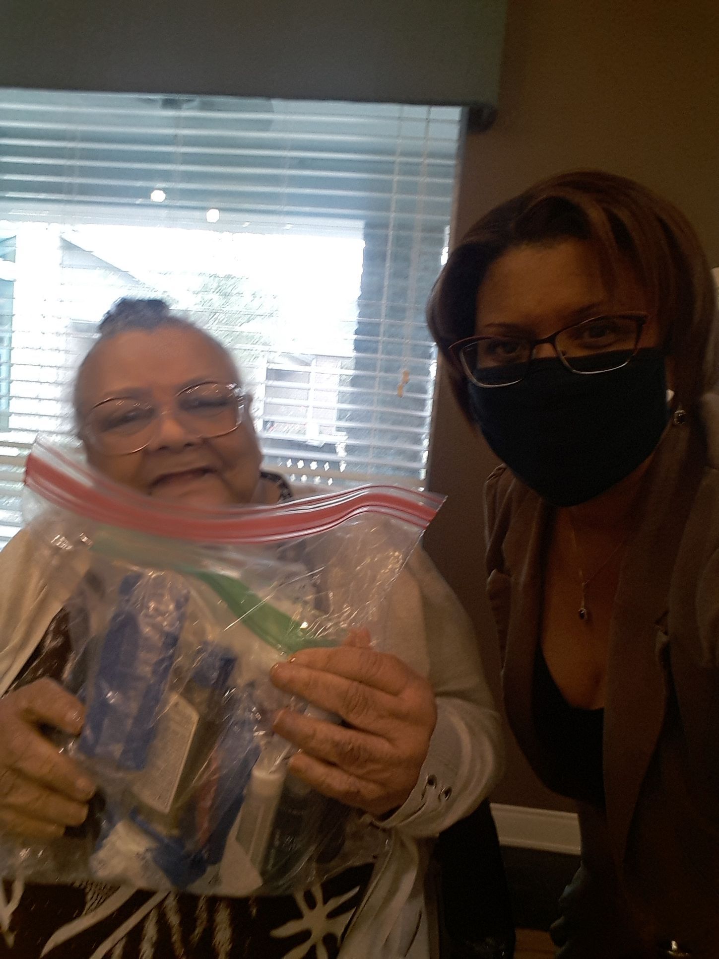 Woman smiles, holding a bag with items; another woman in a mask stands beside her.