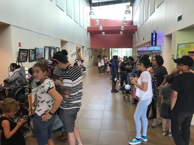 People gather in a building hallway with arcade games, a waiting area, and windows.